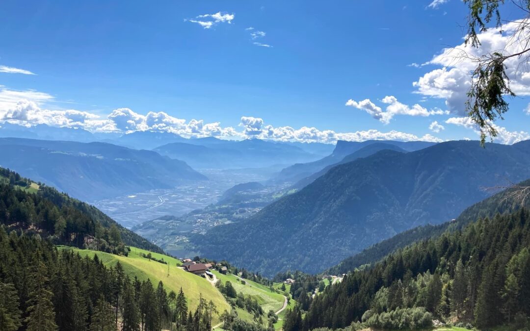 Blick von einem Bergbauernhof oberhalb von Lana in Südtirol über das weite Etschtal und die umliegenden Alpengipfel bei blauem Himmel.