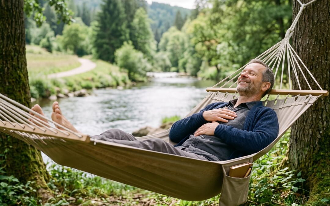Ein entspannter Mann liegt zufrieden und lächelnd mit geschlossenen Augen in einer Hängematte am Ufer eines Flusses in der Natur und genießt den Moment.